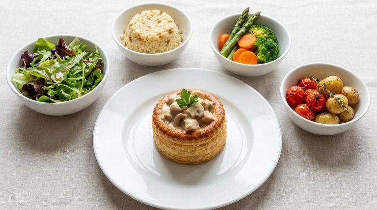 Une bouchée à la reine sur une assiette entourée de bols de salade, riz, légumes vapeur et pommes de terre rôties.