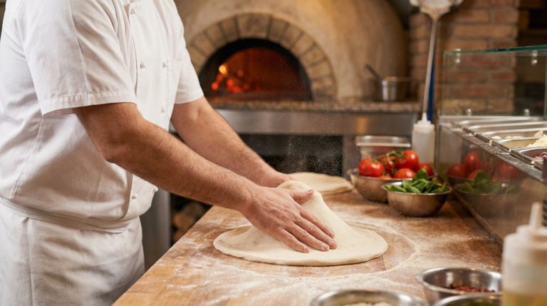 Un pizzaiolo en tenue blanche étalant une pâte à pizza sur un plan de travail en bois devant un four à bois.
